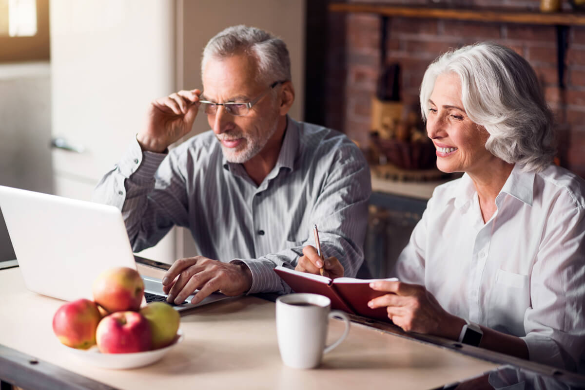 Two older adults, female and male, white hair, female is holding a book the male looking at laptop. They are together in a kitchen setting.