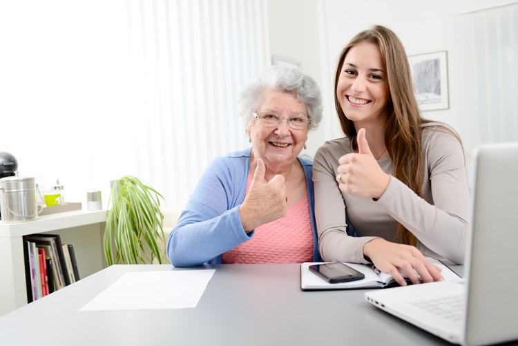 Grandmother and granddaughter giving the thumbs up, sign in front of a laptop with an mobile phone on the table below them